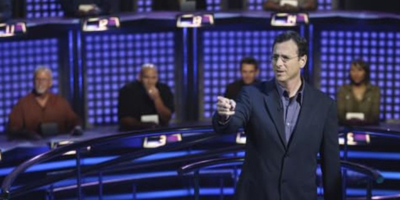 Bob Saget stands on the set in front of contestants and points at something off-screen in 1 vs 100.