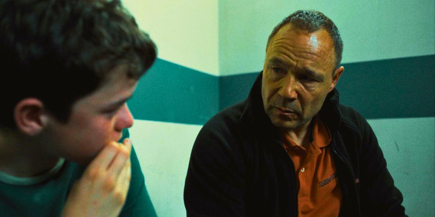 Stephen Graham wears an orange shirt with a black sweater over it and talks to a young boy inside a police station