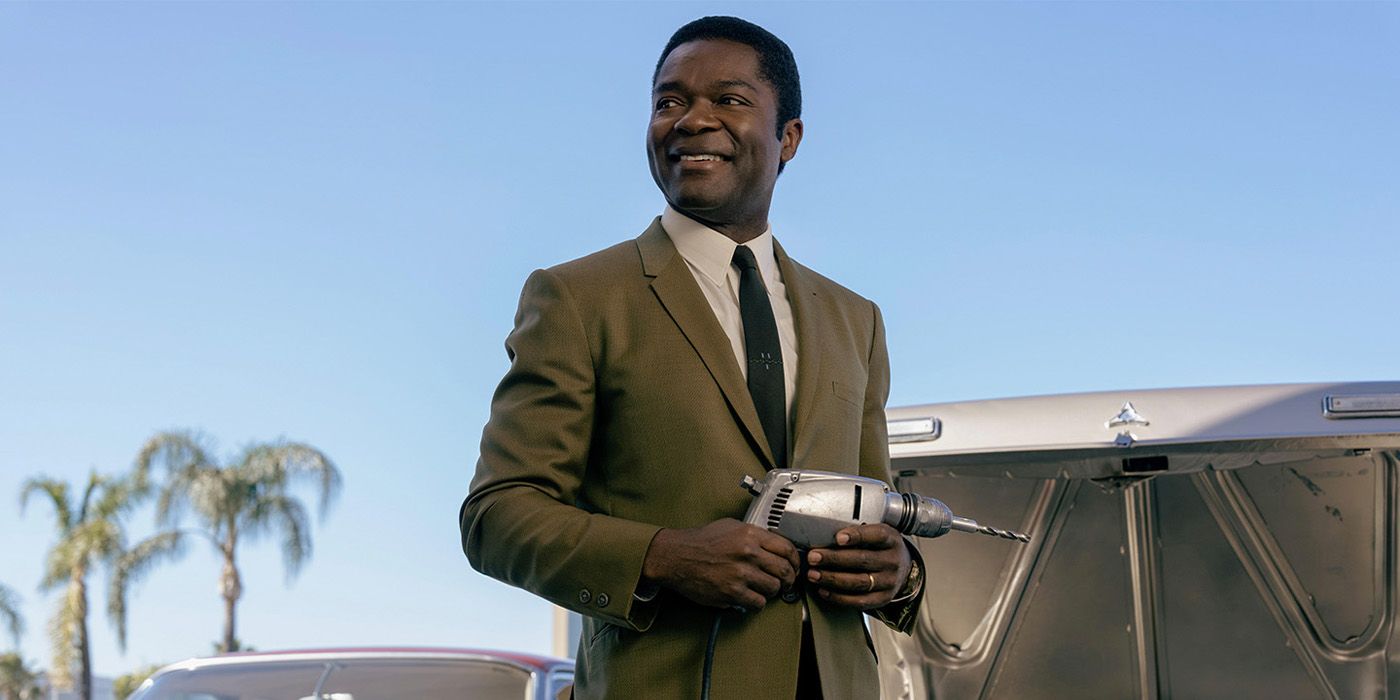 David Oyelowo wearing a suit, smiling, and holding a drill next to a car in front of palm trees