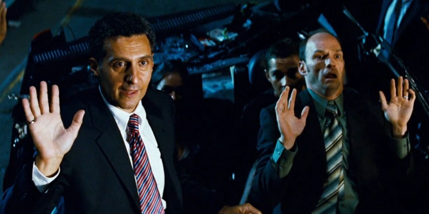 John Turturro and a costar hold up their hands and look surprised with people in a vehicle in Transformers.