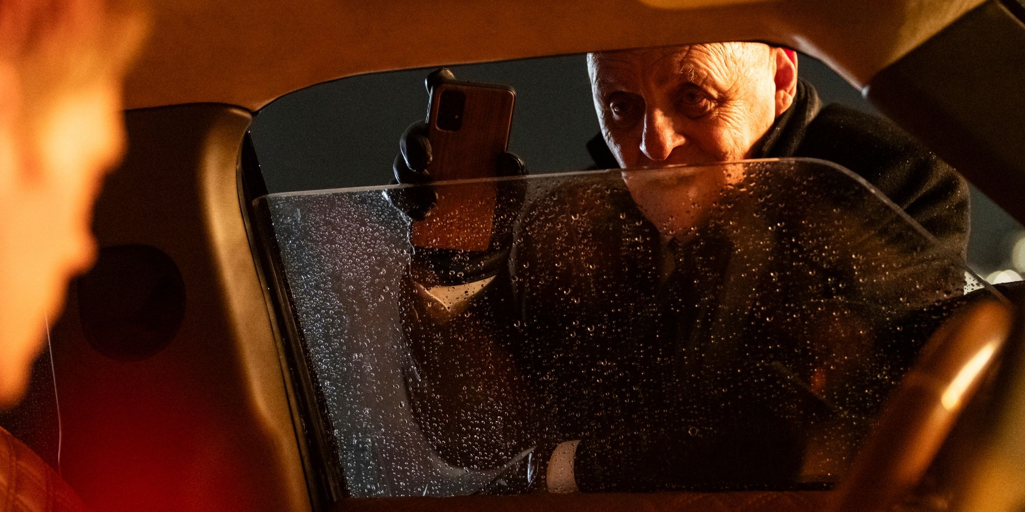 William, played by actor Anthony Hopkins, stands outside a car on a dark, rainy night, holding a cellphone in front of the slightly open window while Eddie, played by actor Bill Skarsgård, looks on from inside in Locked.
