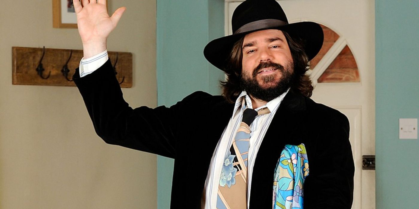 Matt Berry as Beef Galore in House of Fools dressed in a preppy black suit and a black hat, waving at the camera