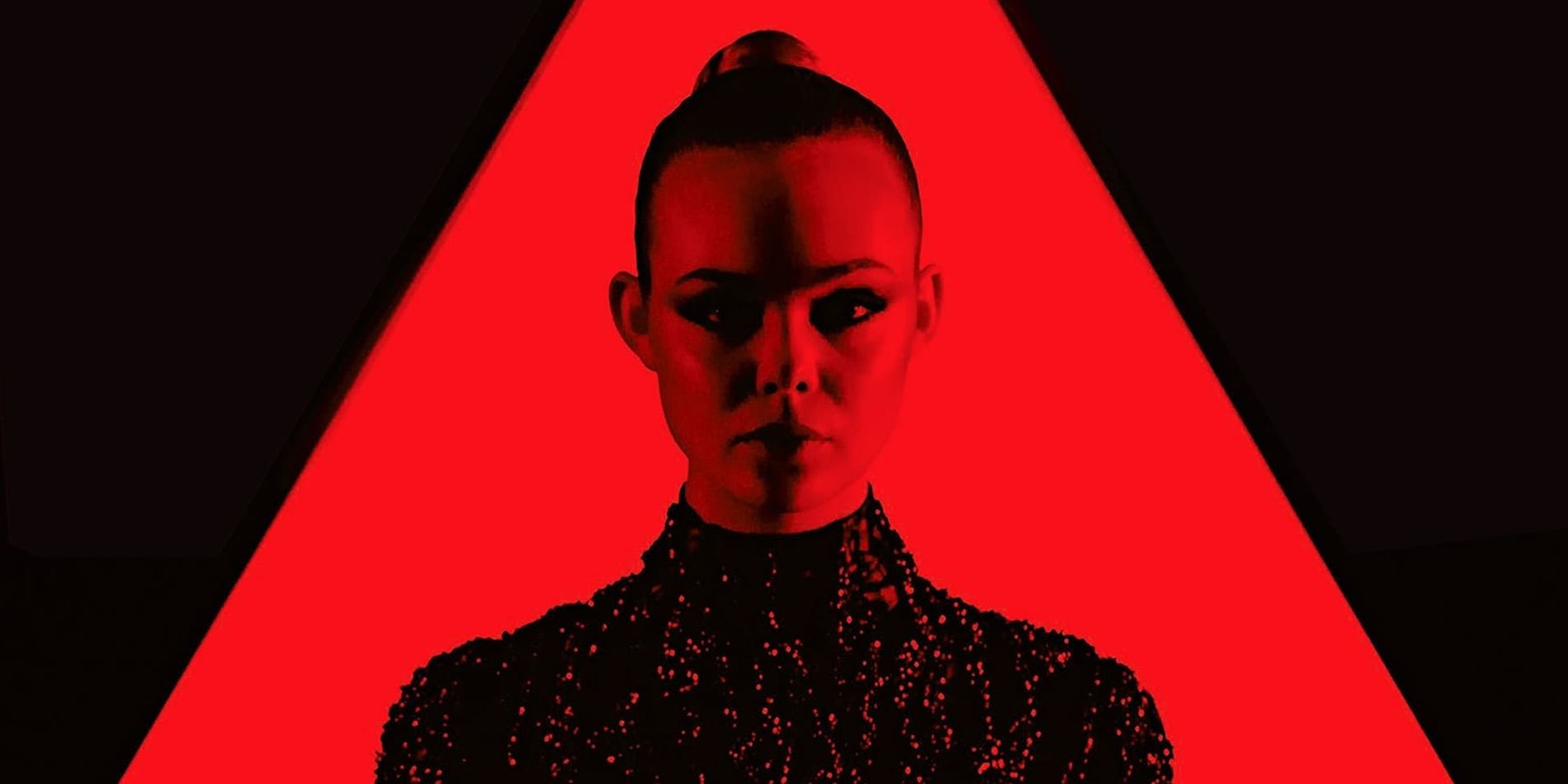 Jesse, played by actor Elle Fanning, stands sternly against a triangular black and red backdrop, her face heavily shadowed, in The Neon Demon