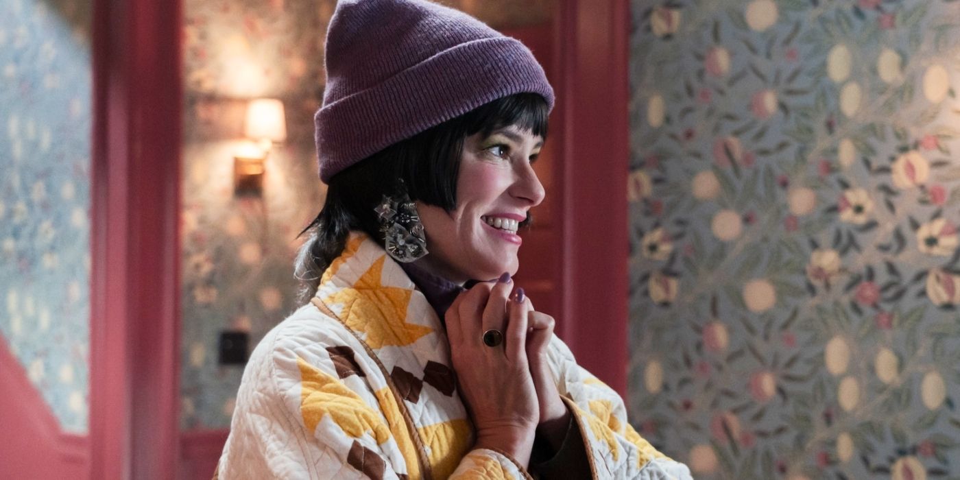 Brenda, played by actor Parker Posey, holds her hands together and grins against a backdrop of floral wallpaper in The Parenting.