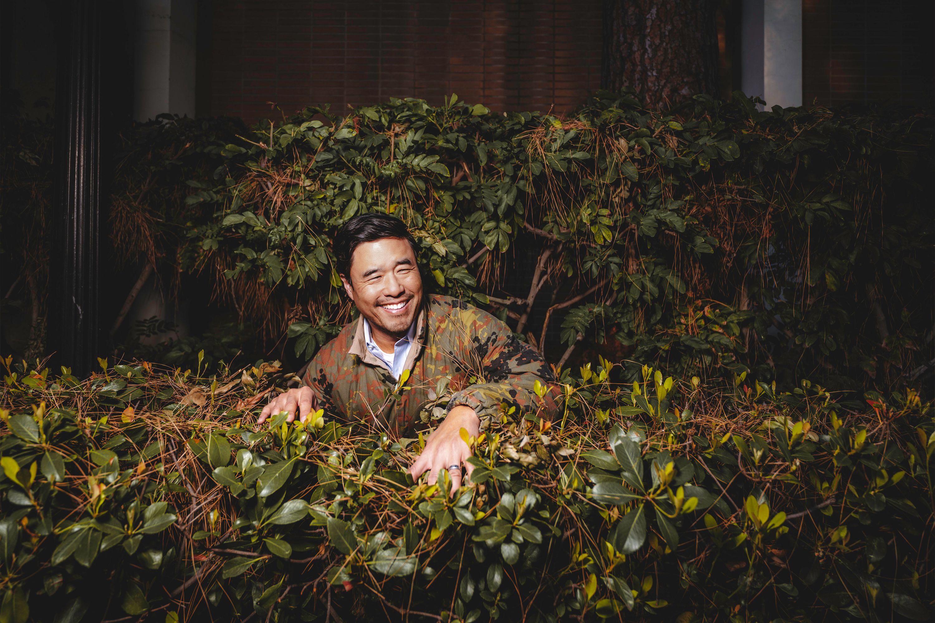 Randall Park wearing a camo jacket while hiding in the bushes at UCLA