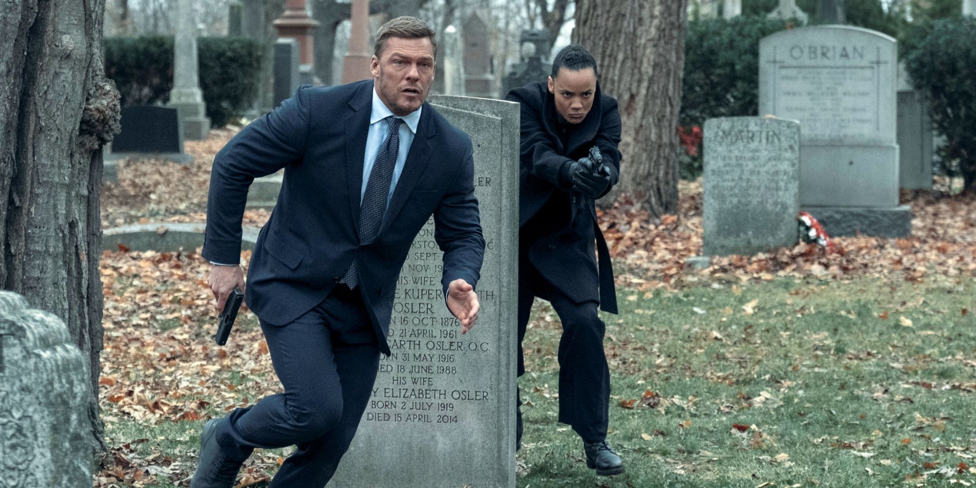 Maria Sten como Neagyy e Alan Ritchson como Jack Reacher correndo por um cemitério com armas em Reacher.