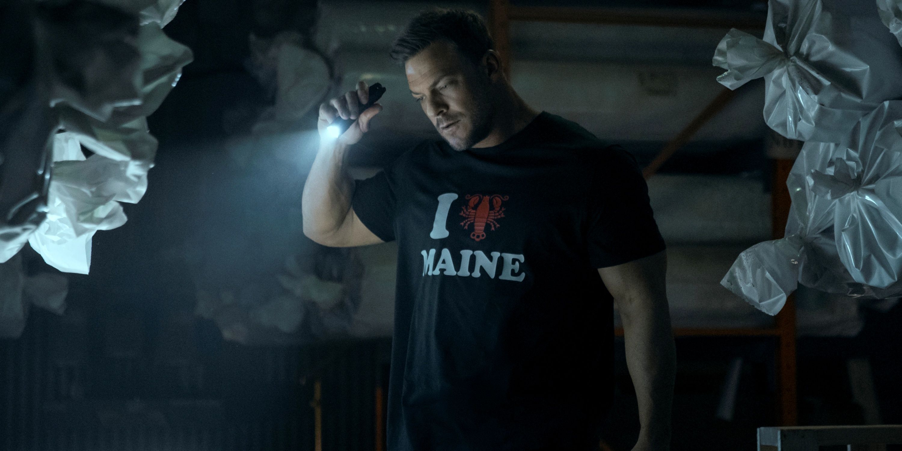 Alan Ritchson as Jack Reacher holding up a flashlight in an &ldquo;I Love Maine&rdquo; t-shirt in Season 3 of Reacher