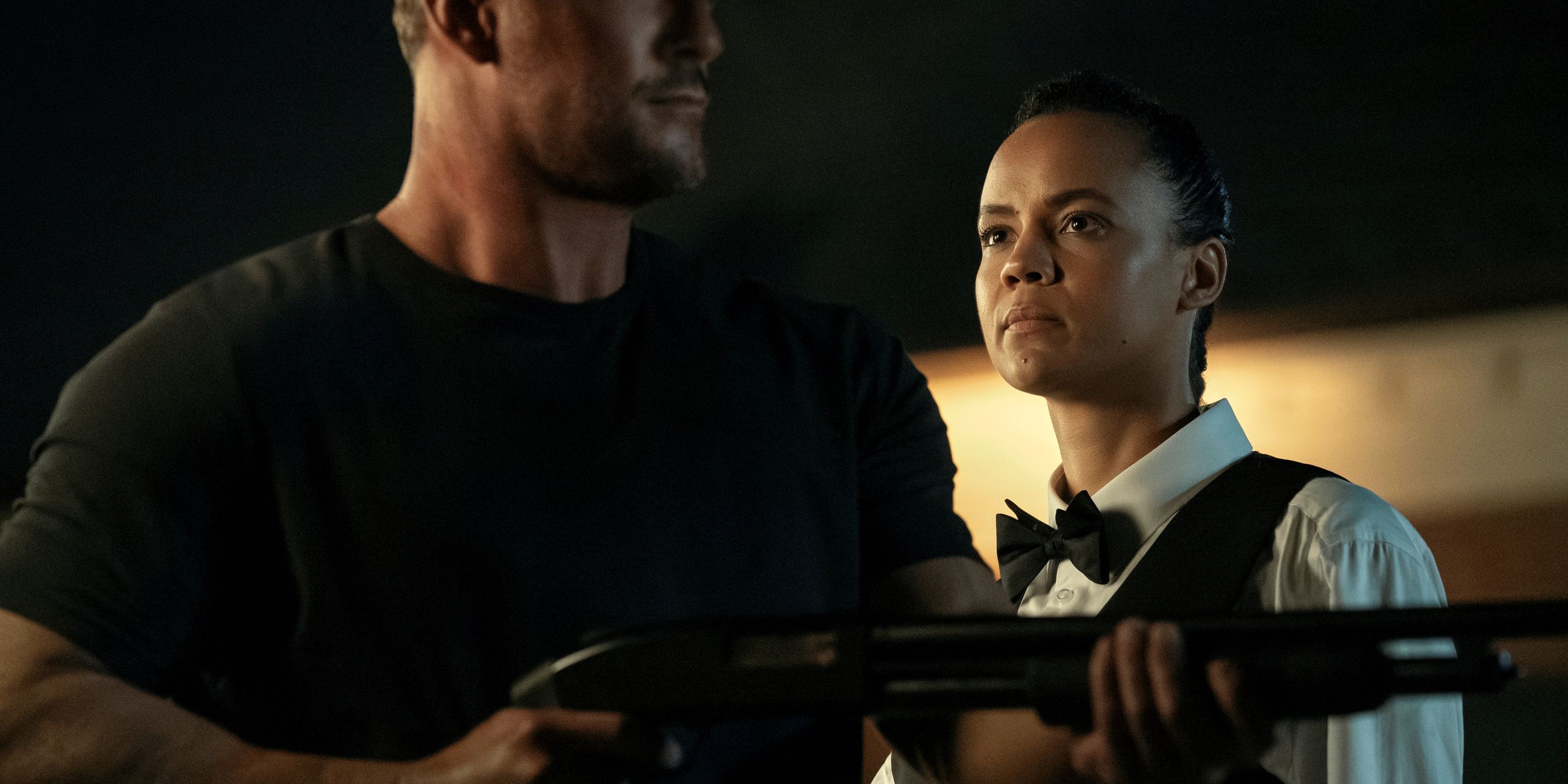 Maria Sten como Neagey, vestida como garçom atrás de Alan Ritchson como Jack Reacher segurando uma arma no Reacher.