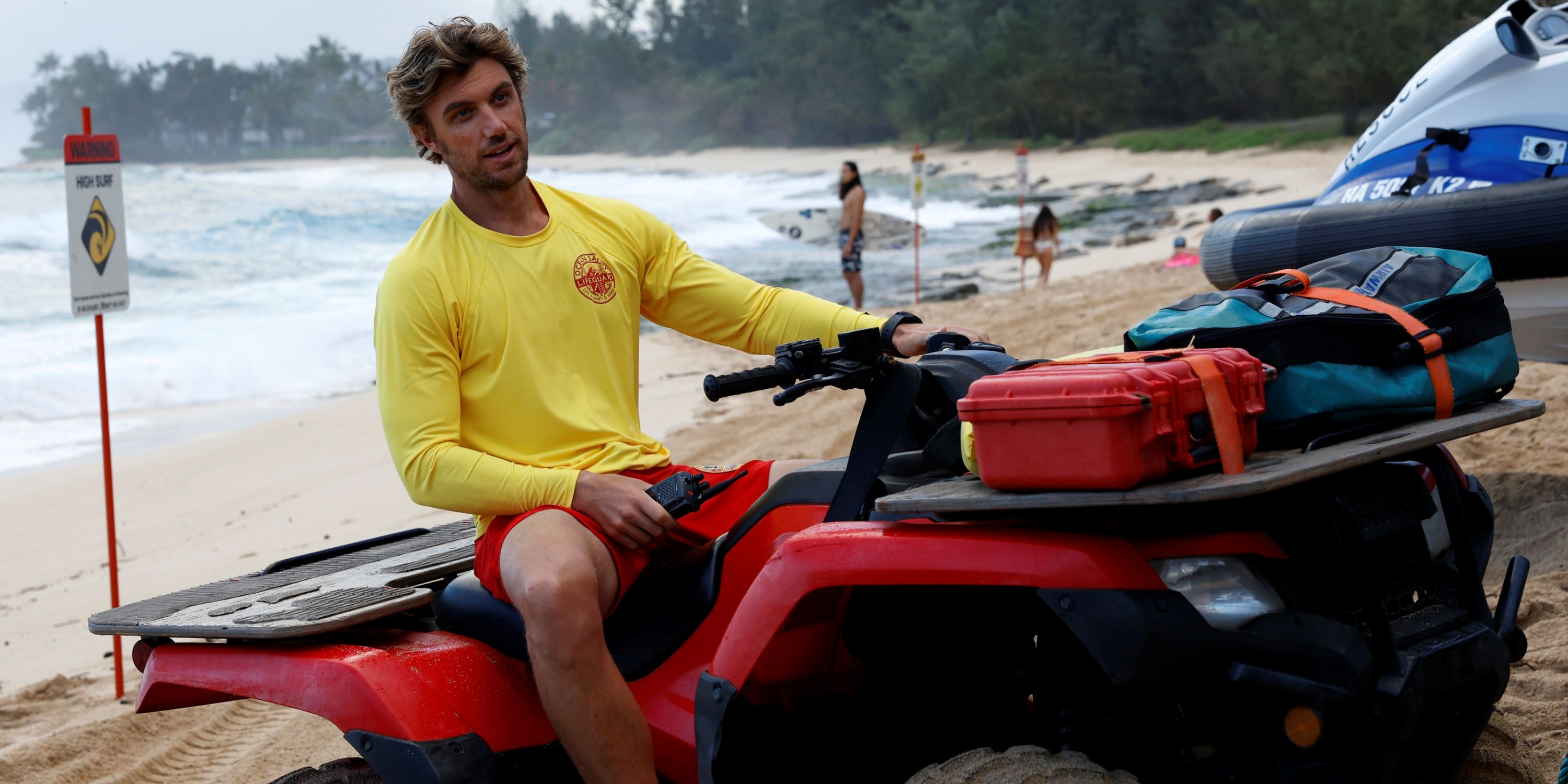 Adam Demos as Will Ready in his yellow lifeguard shirt and red shorts on a beach vehicle in Rescue: HI-Surf