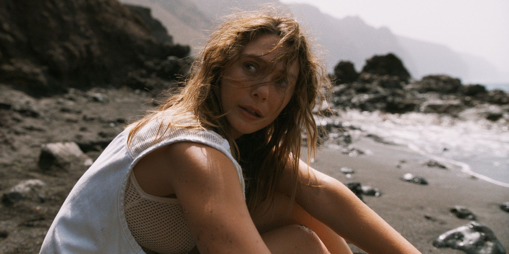Elizbeth Olsen as Mia sitting in the sand on the beach and looking over her right shoulder in The Assessment