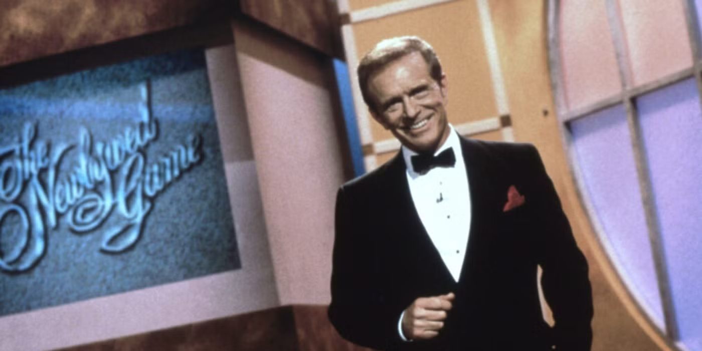 Bob Eubanks, the host of The Newlywed Game stands on set and smiles.