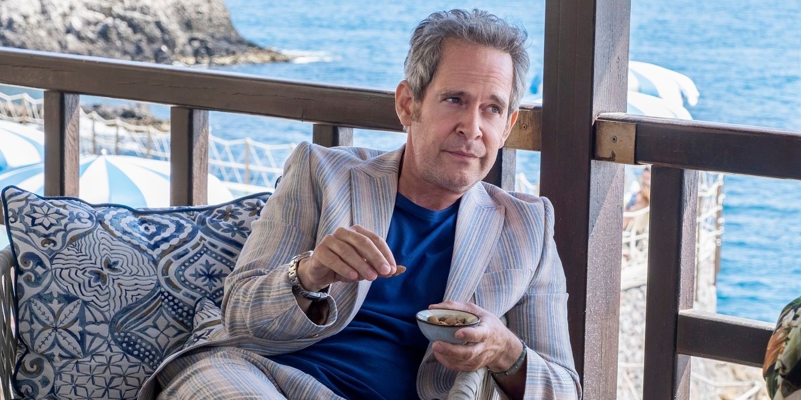 Tom Hollander as Quentin holding a small bowl on a balcony above water in The White Lotus.