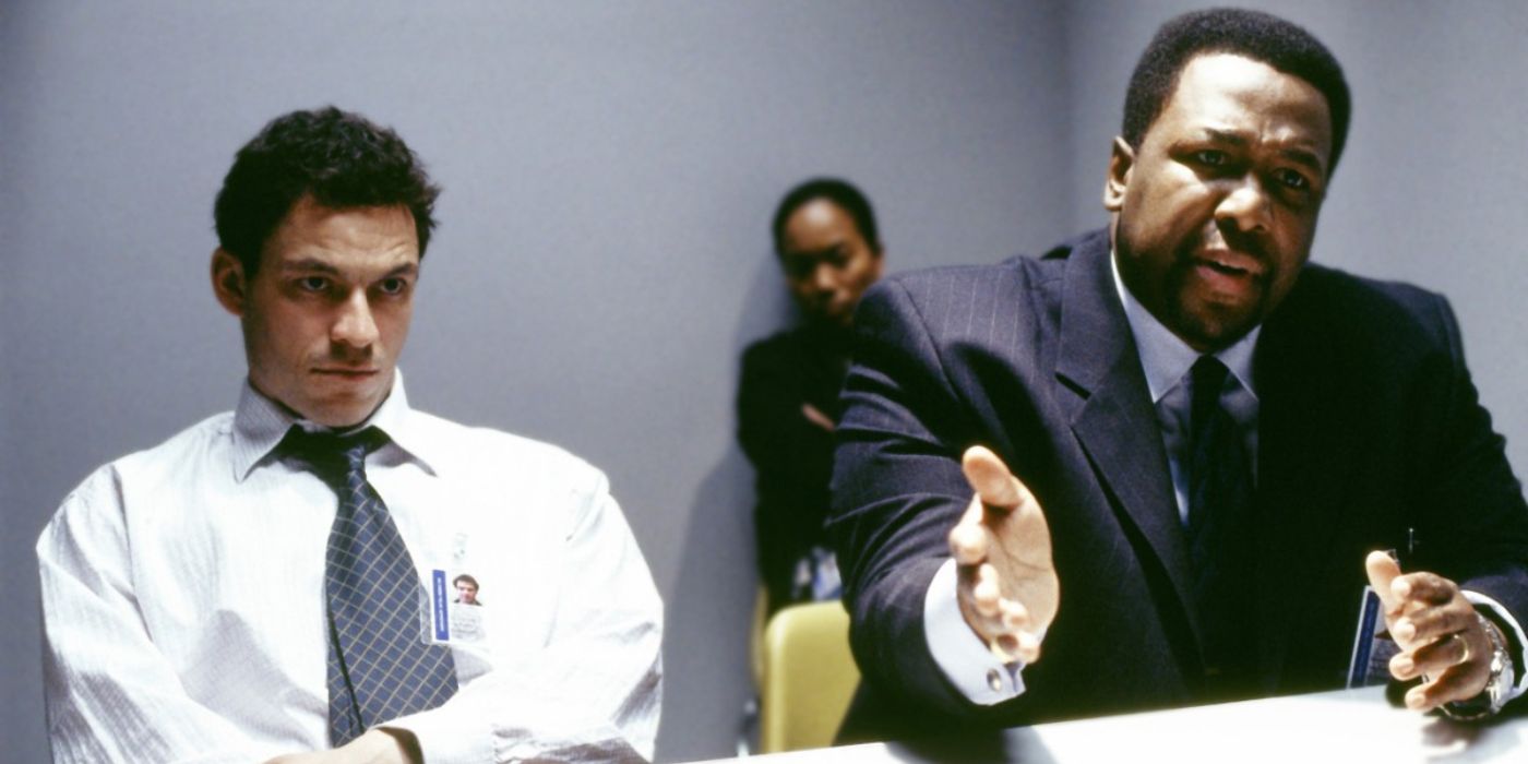 McNulty and Bunk sit at a table in an interrogation room with a woman sitting in the background in The Wire.
