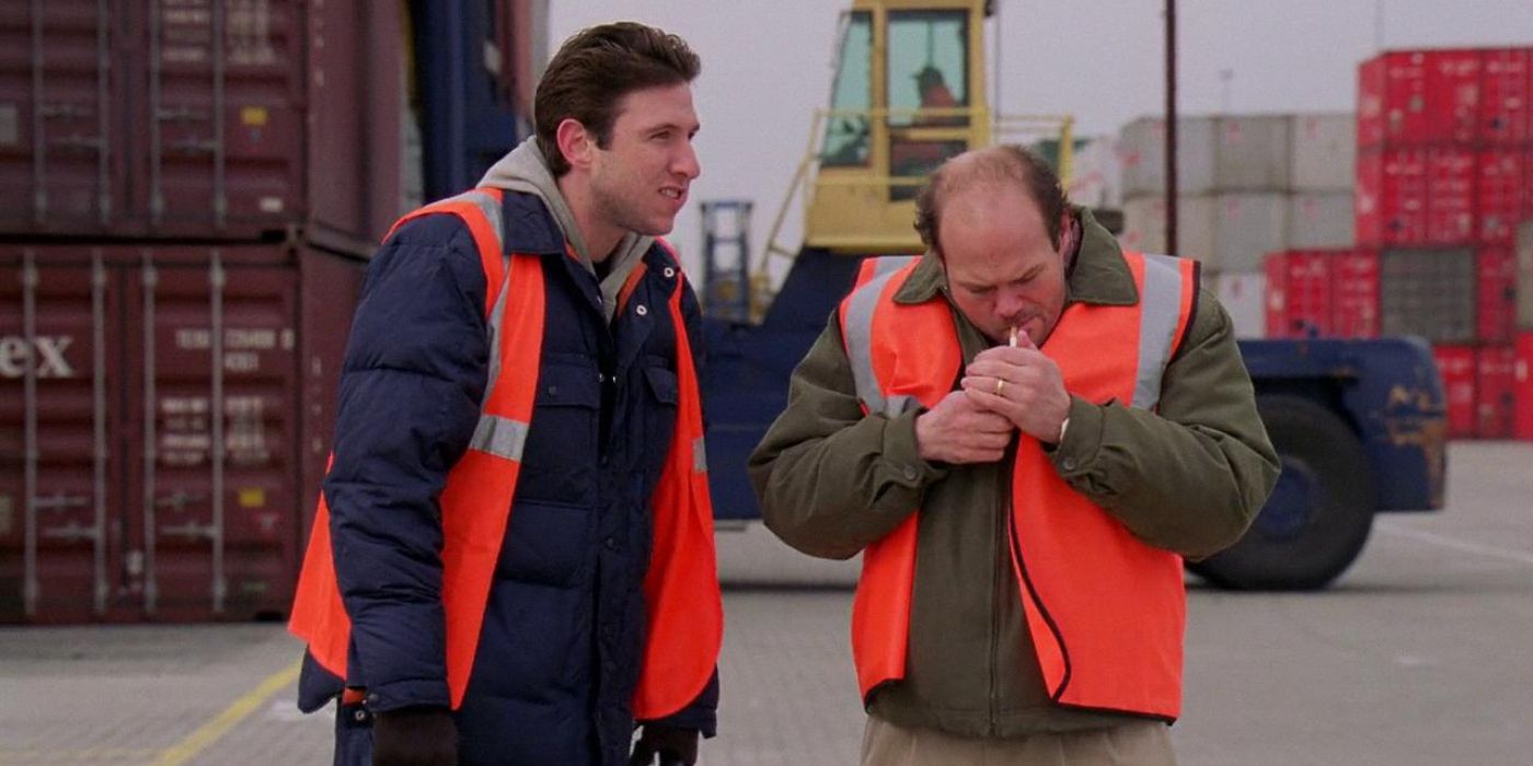 One man lights a cigarette as another stands near, both in bright orange vests in a shipyard in The Wire.