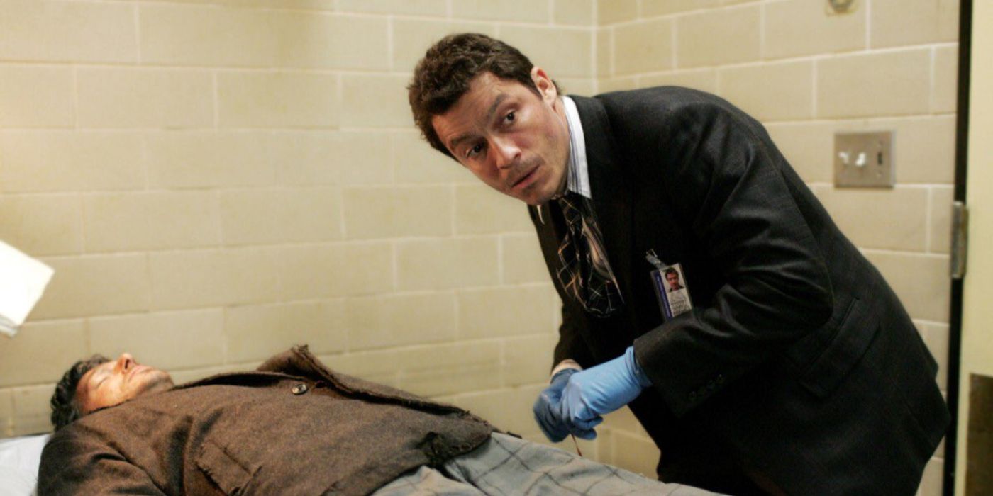 A man with sterile gloves on looks over his shoulder, leaning over a body on a table in The Wire.