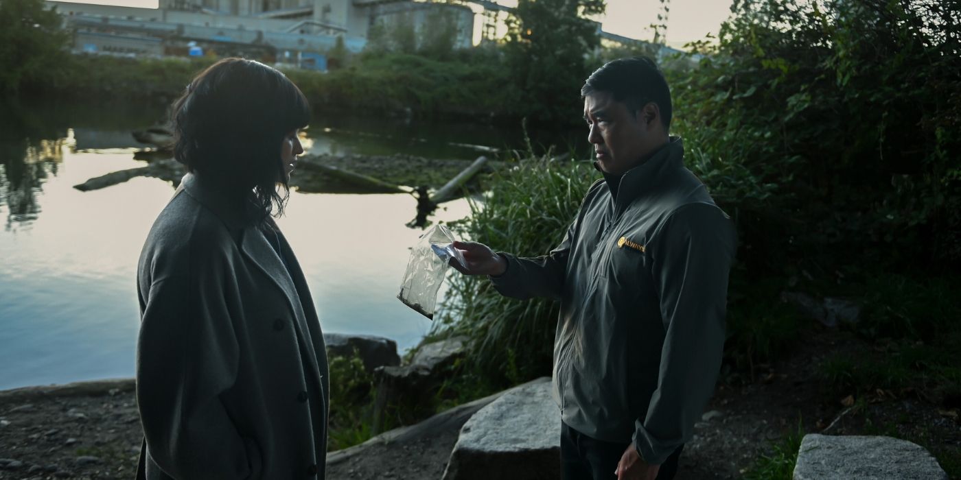 Eve Hollow and Randall Park as Ingrid Derian and Moriarty, standing by a pond while Moriarty holds out a small bag in Watson Episode 7