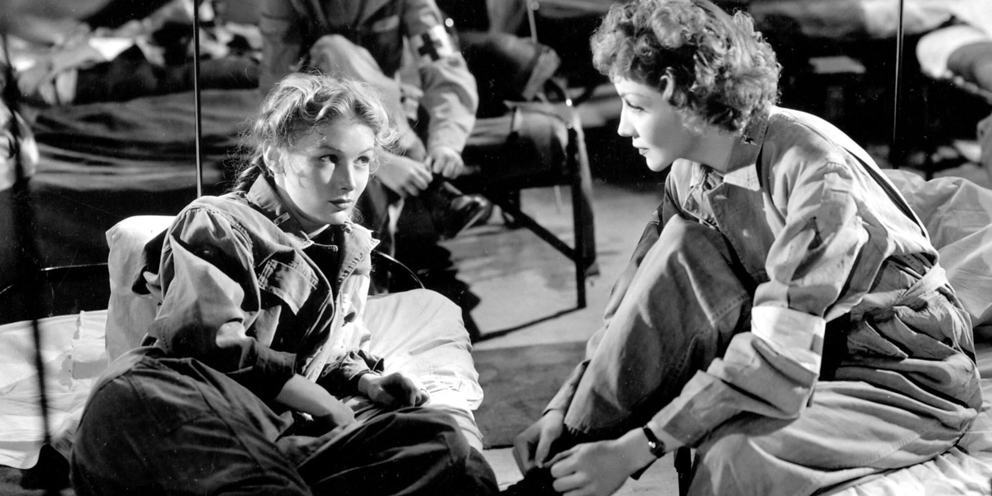 Veronica Lake on a cot looks at Claudette Colbert, sitting near on another cot in So Proudly We Hail, 1943.