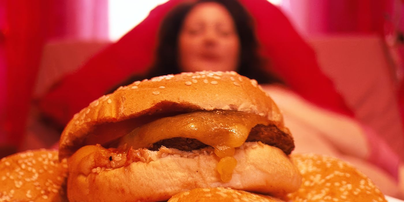 A pile of cheeseburgers in the foreground, with Deidre, played by actor Gabby Millgate, lying in the background looking on in Feed.