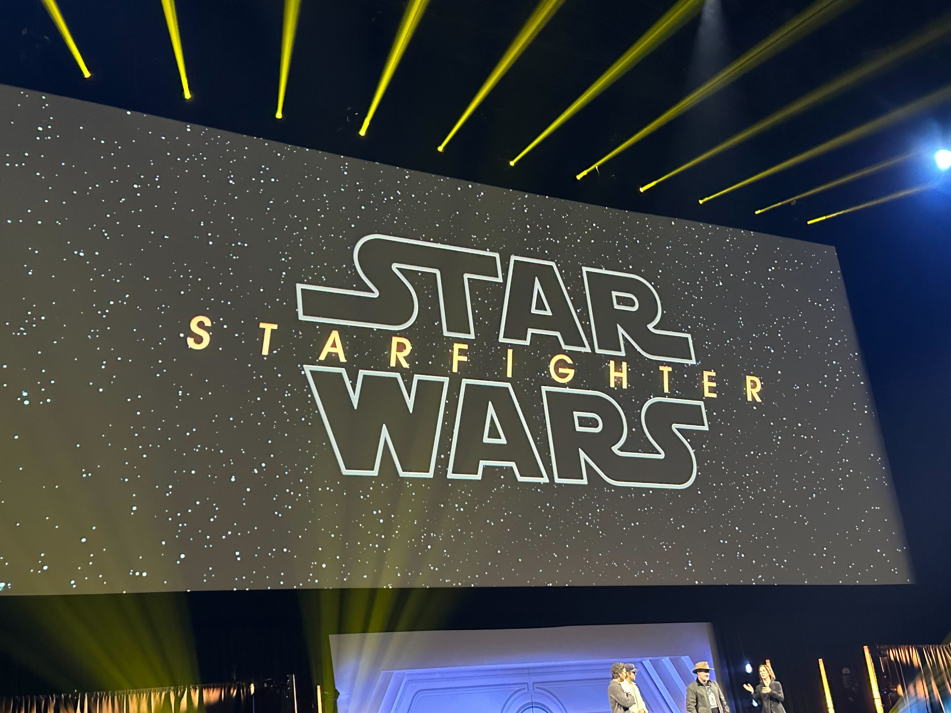 A projection screen with the title image for Star Wars Starfighter with people standing on the stage below