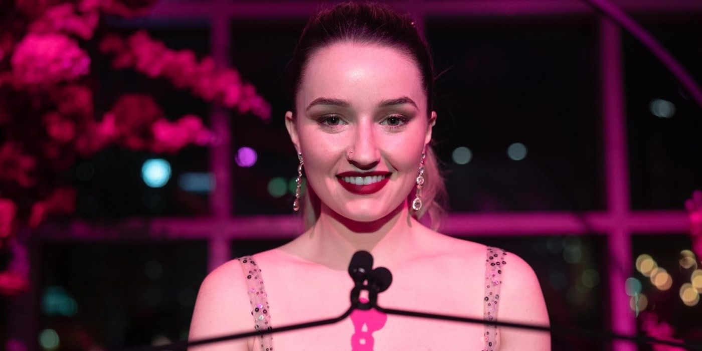 Kaitlyn Dever standing in front of a microphone and smiling in an evening dress in Apple Cider Vinegar.