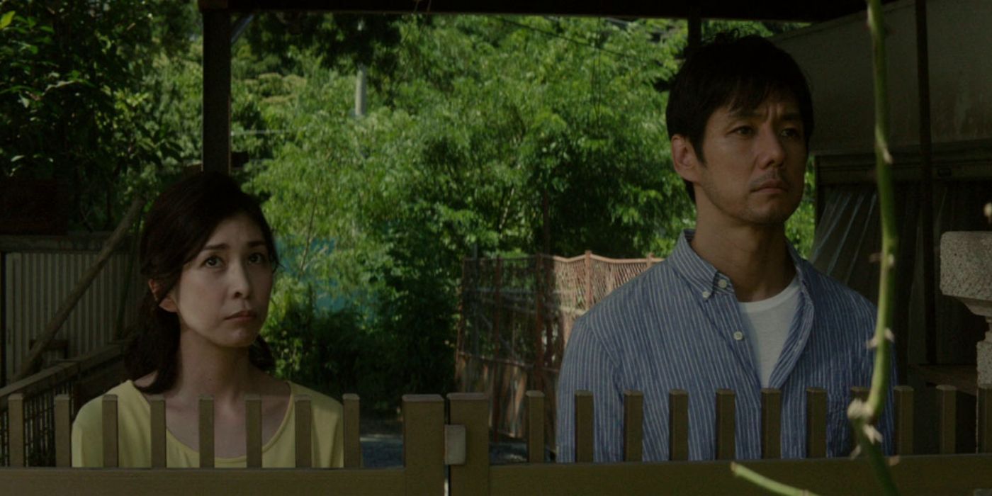 Koichi (Hidetoshi Nishijima) and Yasuko (Yūko Takeuchi) standing next to a fence and watching something off-screen in Creepy