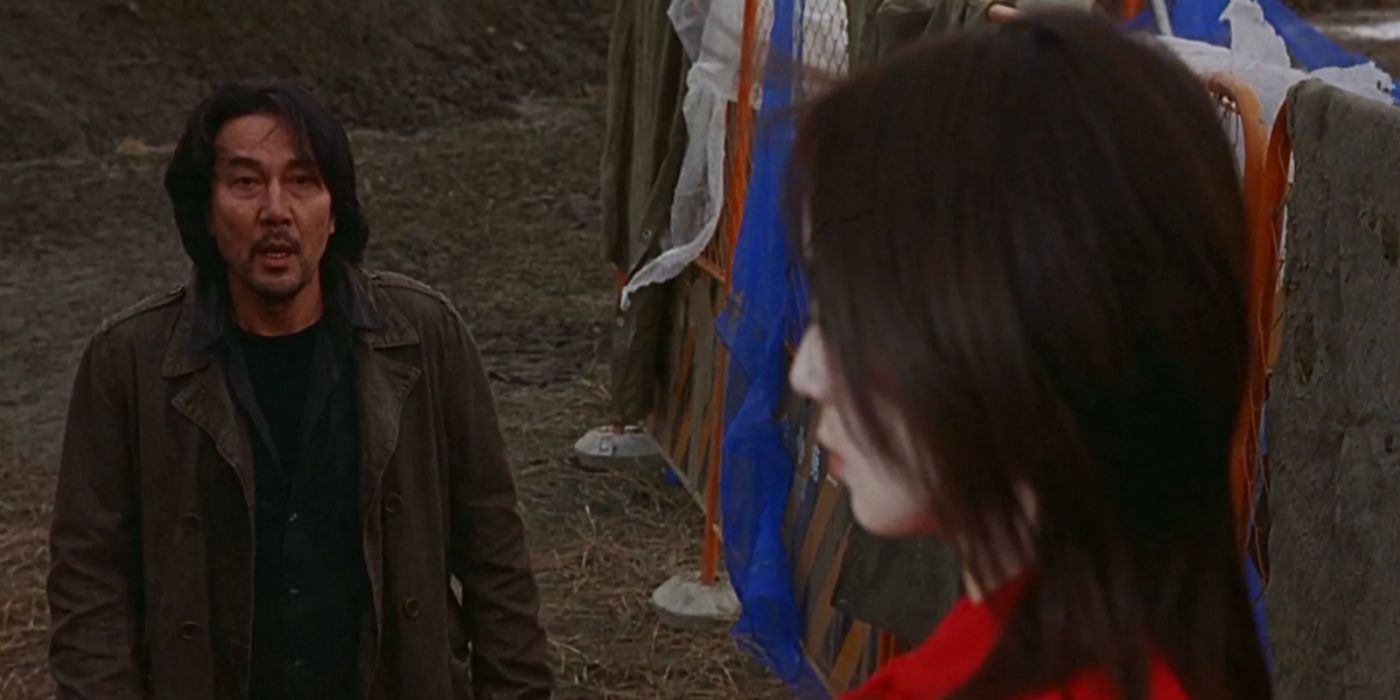 Koji Yakusho looking at Riona Hazuki wearing red and standing closer to the camera with her left cheek turned in Retribution