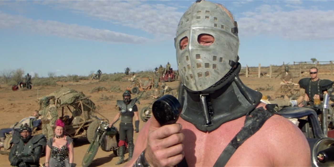 A person with a metal covering over their face with others and makeshift cars in Mad Max 2 The Road Warrior.