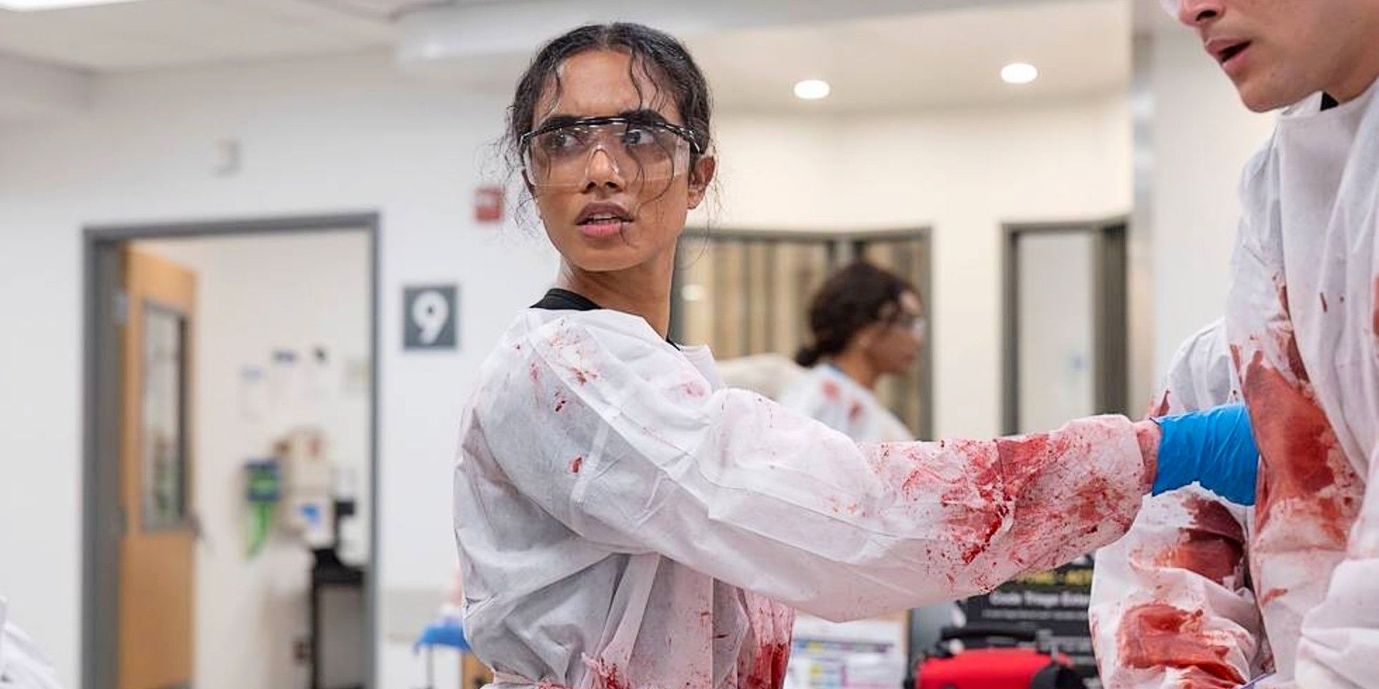 Mohan in bloody scrubs and safety glasses treating a patient in The Pitt Season 1.
