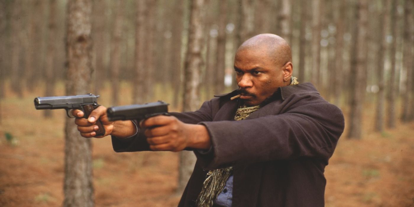 Ving Rhames as Mann holding guns in both hands and aiming at something off-camera in a forest in Rosewood.
