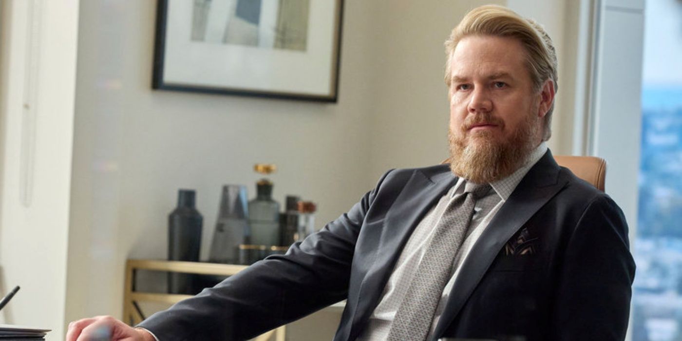 Josh McDermitt as Stuart Lane sitting in a chair at his office desk with his right arm extended out in Suits LA
