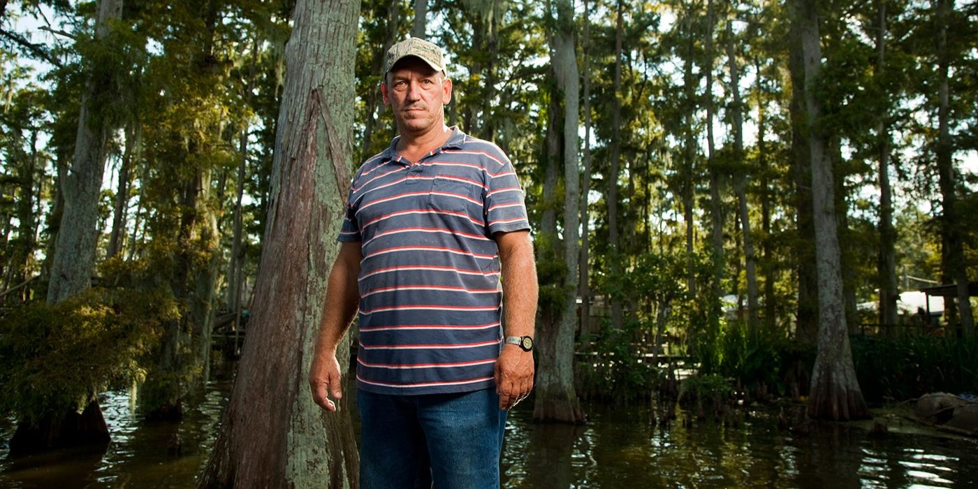 Troy Landry standing outside with trees and water in a striped shirt in a promo photo for Swamp People.