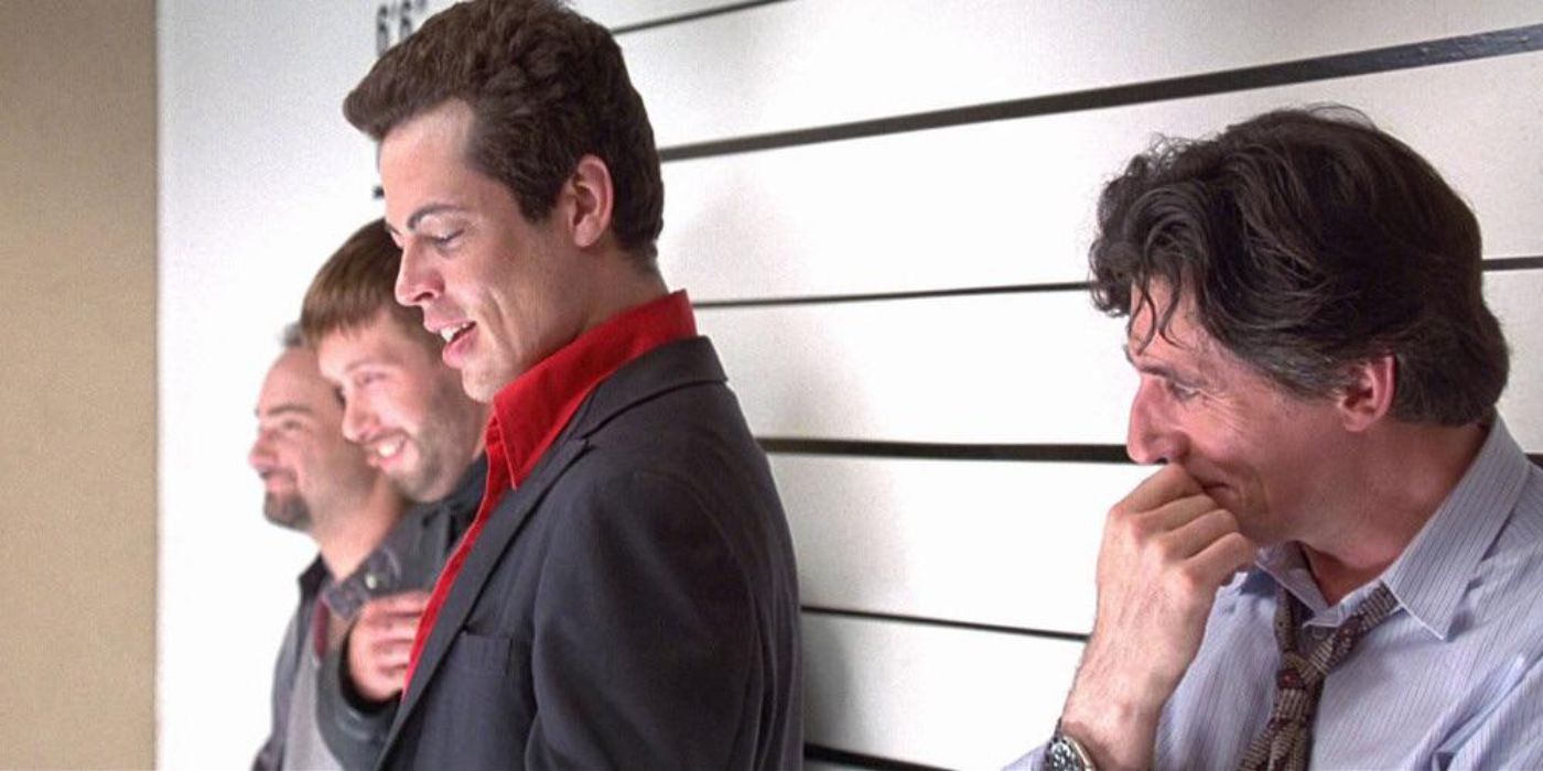 Kevin Pollack, Stephen Baldwin, and Gabriel Byrne leaning on a lineup wall with Benicio del Toro reading a card and smiling in The Usual Suspects