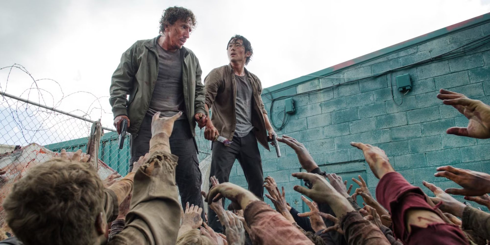 Steven Yeun as Glenn and Michael Traynor as Nicholas stand on a bin as zombies reach up in The Walking Dead.