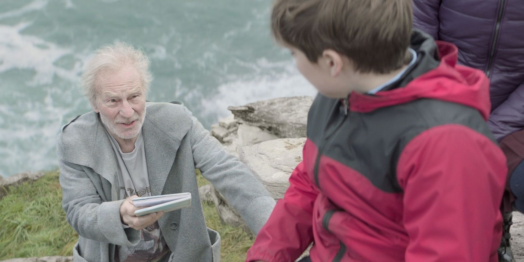 Patrick Stewart as Merlin hands a notebook to a child while on a cliffside in The Kid Who Would Be King, 2019.