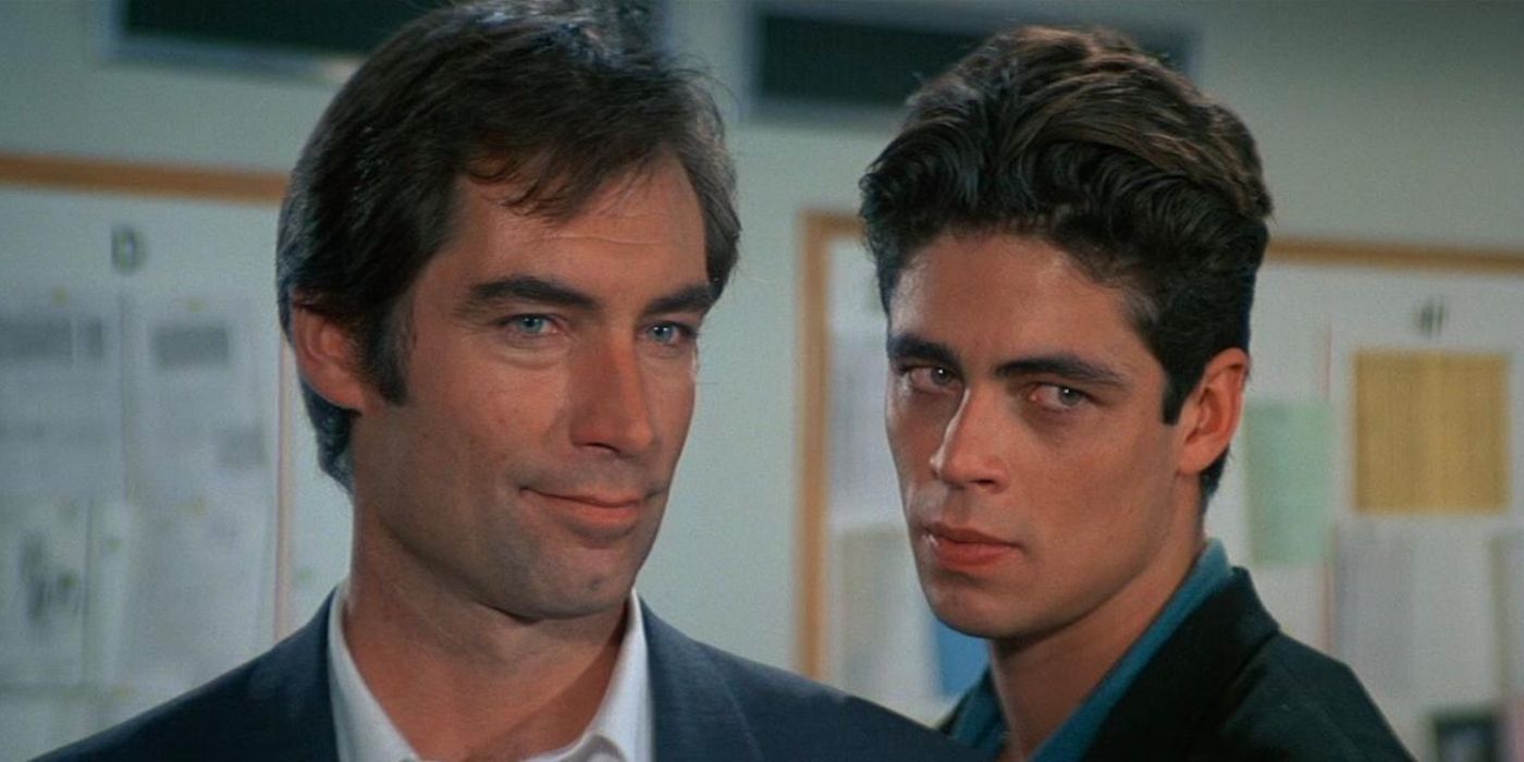 Benicio del Toro em pé atrás de Timothy Dalton e sorrindo em licença para matar