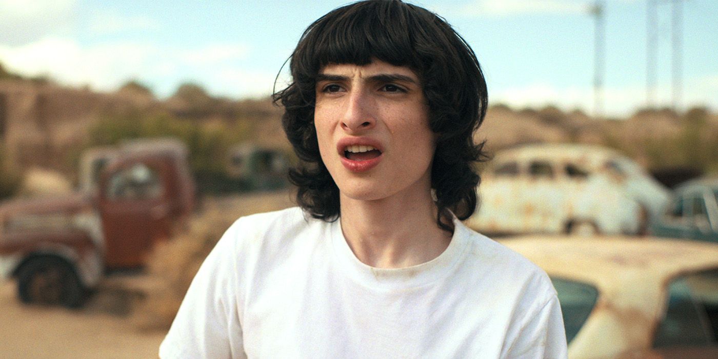 Mike Wheeler (Finn Wolfhard) looking confused, standing in a junkyard with rusty cars in Stranger Things.