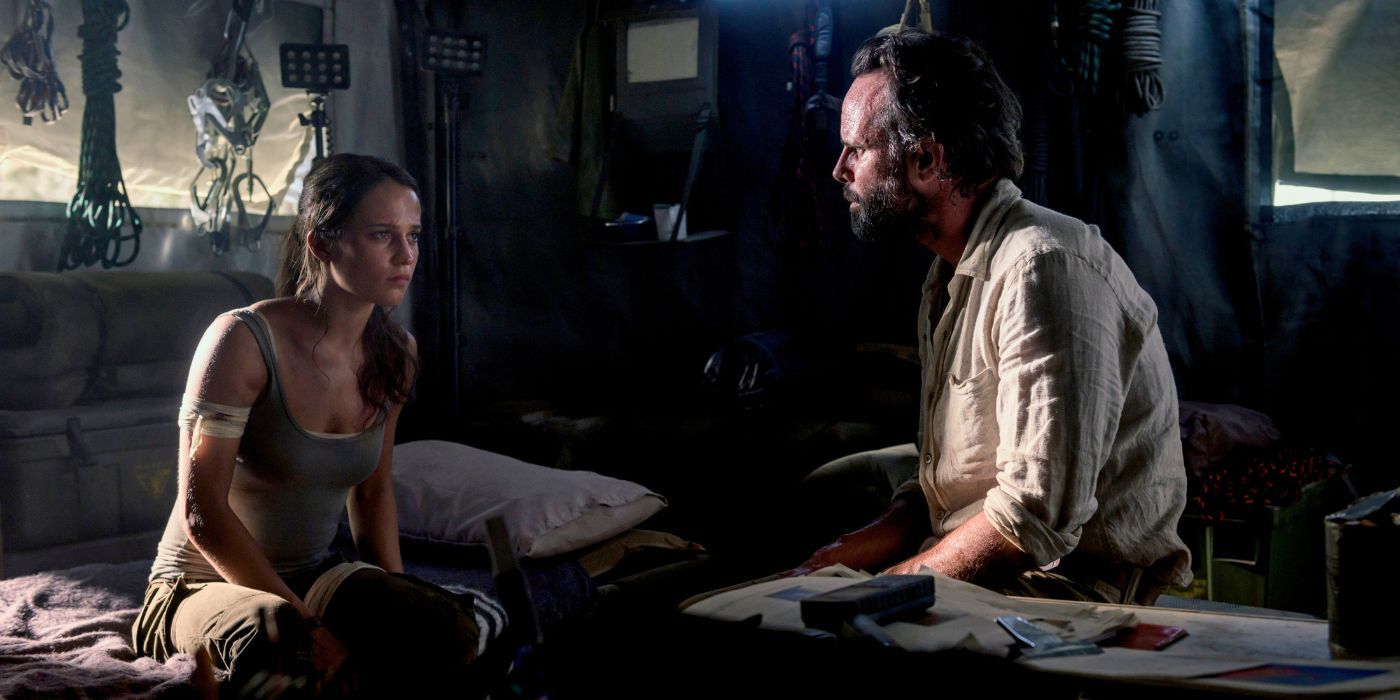 Alicia Vikander como Lara Croft sentada em um acampamento conversando com Walton Goggins como Mathias Vogel em Tomb Raider.