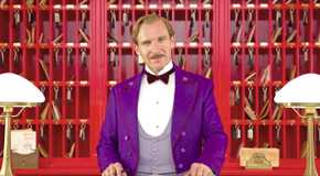 Ralph Fiennes as M. Gustave in a purple tuxedo stands behind the front desk in The Grand Budapest Hotel.