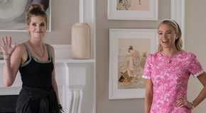 Meghann Fahy waving wearing a black tank top standing next to Milly Alcock smiling wearing a pink dress with one hand on her hip