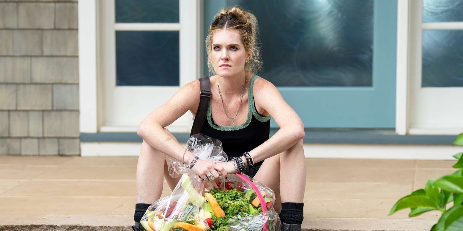 Meghann Fahy wearing a black tank top and boots, sitting on the steps while holding a fruit basket in Sirens