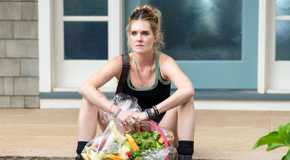 Meghann Fahy wearing a black tank top and boots, sitting on the steps while holding a fruit basket in Sirens