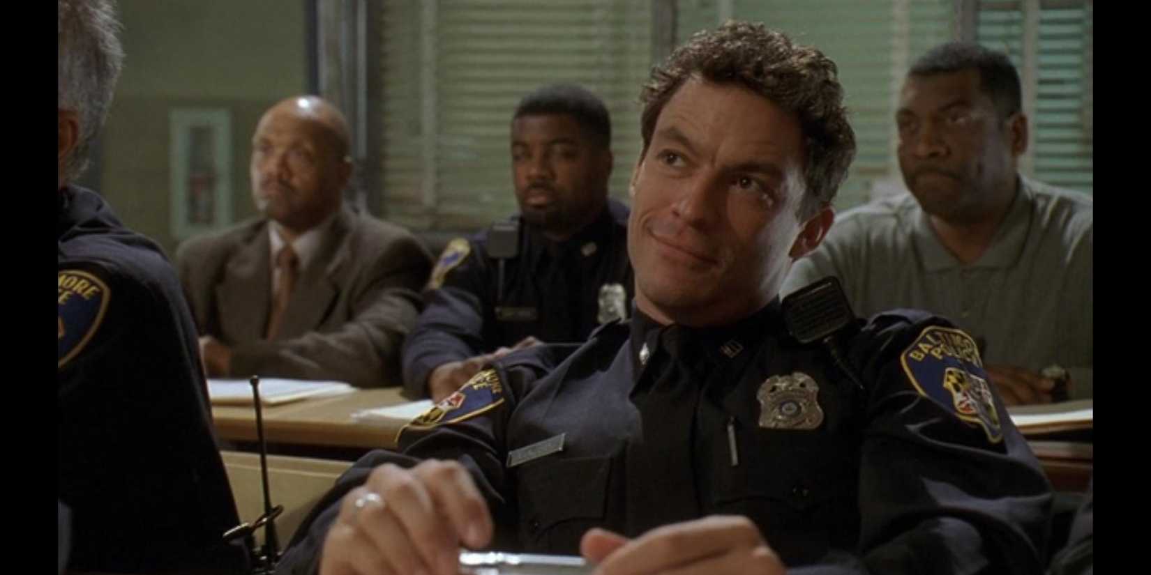 Dominic West in uniform sits at a desk in a room with others in the Mission Accomplished episode of The Wire.