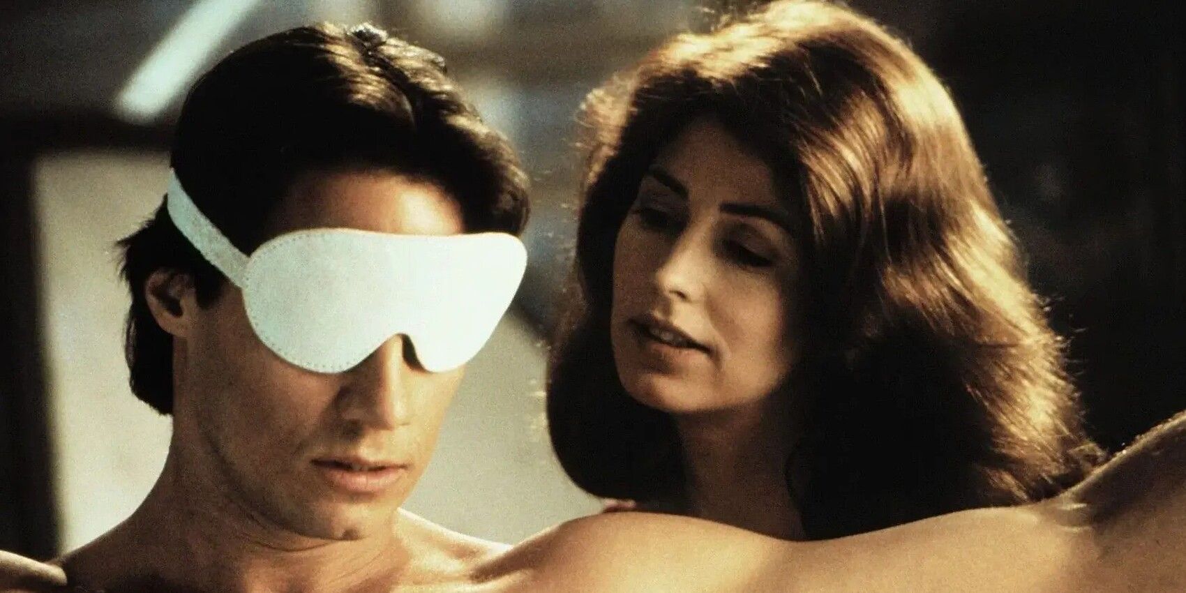 A woman looking over the shoulder of a shirtless man wearing a white eyemask in 'Exit to Eden'