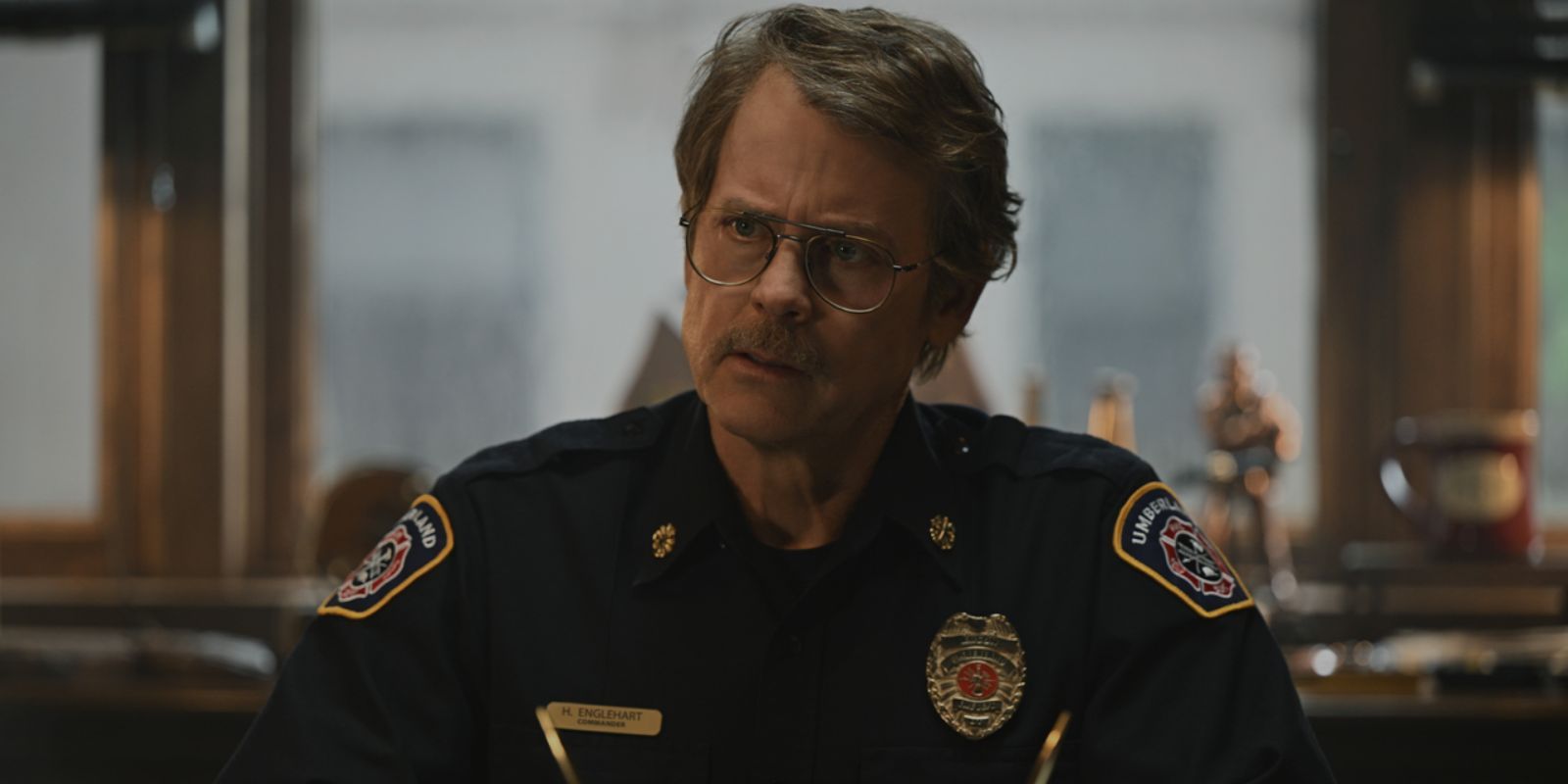 Greg Kinner franzindo a testa e olhando atentamente para alguém fora da câmera em fumaça