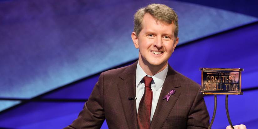 Ken Jennings on the 'Jeopardy!' stage, smiling and holding a trophy that says "Greatest of All Time."