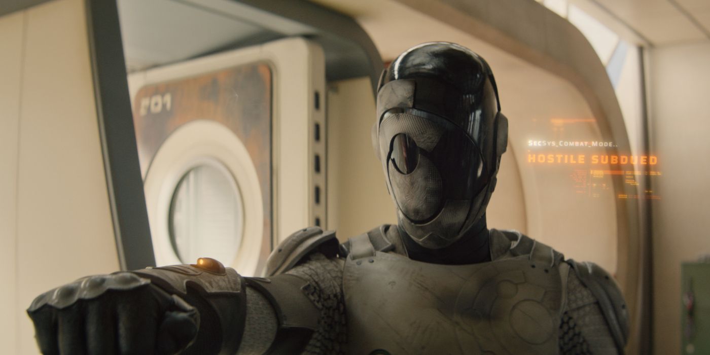 Murderbot with its helmet up and aiming its arm gun