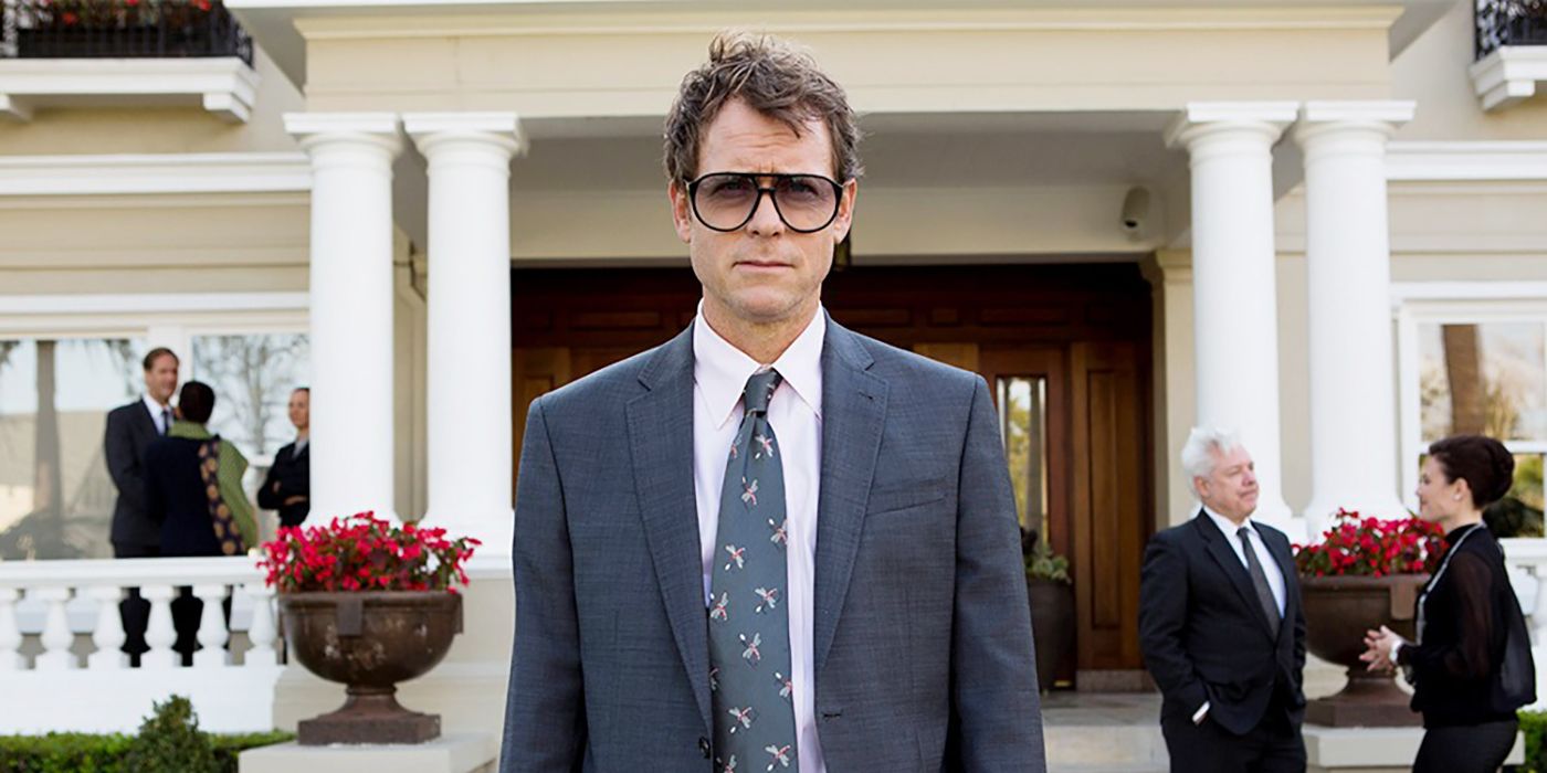 Greg Kinnear em pé em frente a um prédio em um terno e óculos de sol em ancinho.