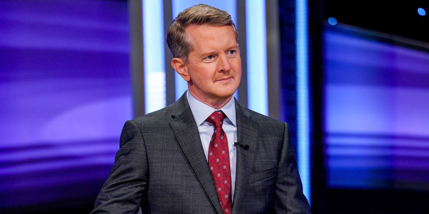 'Jeopardy!' host Ken Jennings standing at the podium, wearing a red tie and a gray jacket.
