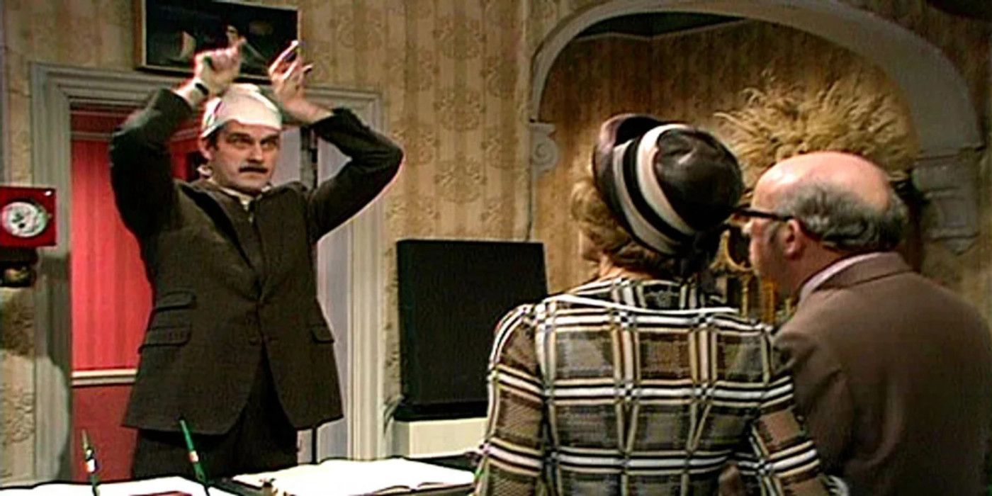 Basil holds his hands above his head at hotel guests in Fawlty Towers.