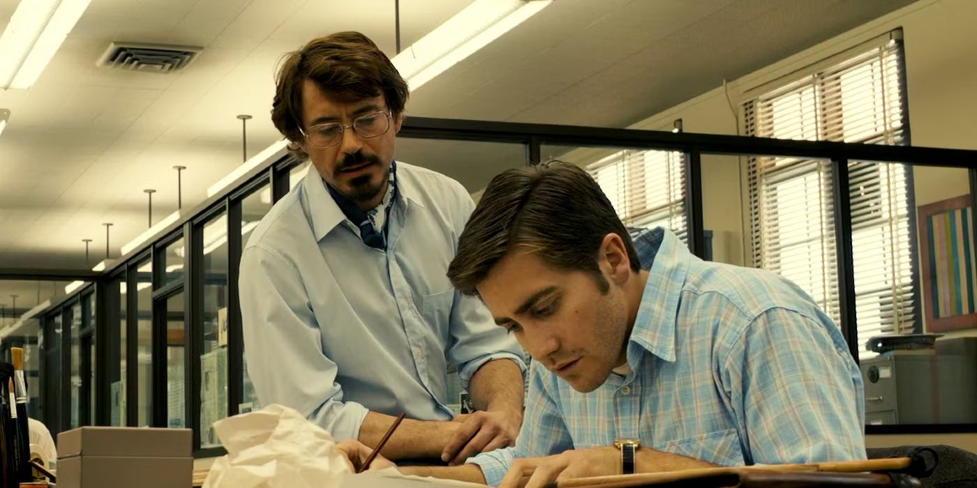 Robert Graysmith (Jake Gyllenhaal) hunches obsessively over his desk while Paul Avery (Robert Downey Jr.) loiters casually behind him in 'Zodiac' (2007).